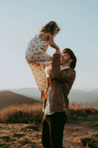 family photography, young daughter climbs up on dad as he holds her hands in the outdoors